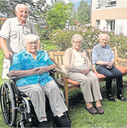Vier ältere Menschen sitzen im Garten eines Pflegeheims – drei auf einer Holzbank, eine Person im Rollstuhl. Ein weiterer älterer Mann steht hinter der Gruppe. Die Szene wirkt freundlich und entspannt an einem sonnigen Tag im Grünen.